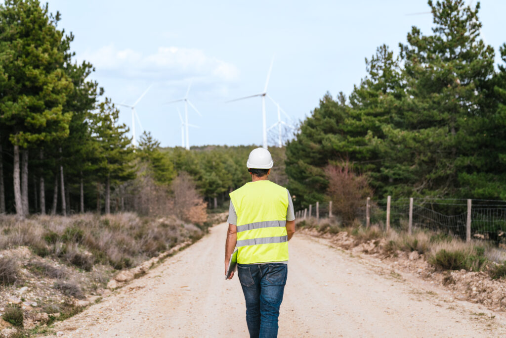 Technician Walking Down Forest Path with Wind Turbines in Background